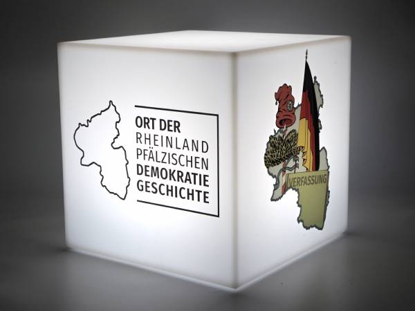 Leuchtender Sitzwürfel mit Aufdruck „Ort der rheinland-pfälzischen Demokratiegeschichte“ und festlichen Motiven.