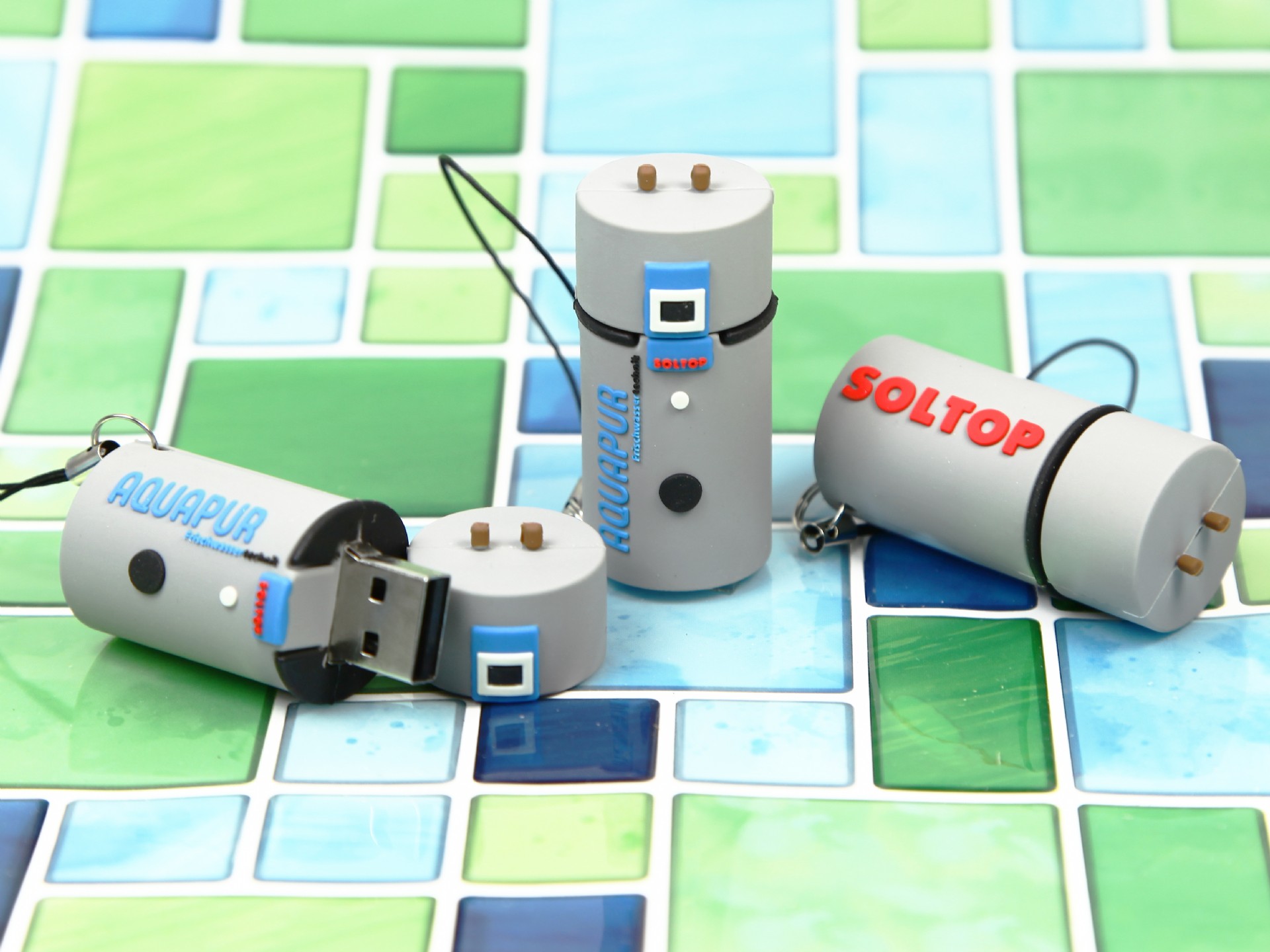 Drei USB-Sticks in Form von Heizungen, bedruckt mit den Logos AQUAPUR und SOLTOP, auf buntem Fliesenhintergrund.