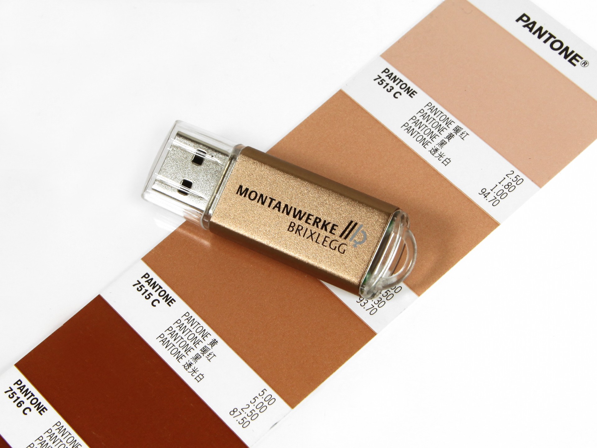 USB-Stick in Gold mit individueller Beschriftung und PANTONE-Farbmustern im Hintergrund. Ideal für Werbezwecke.