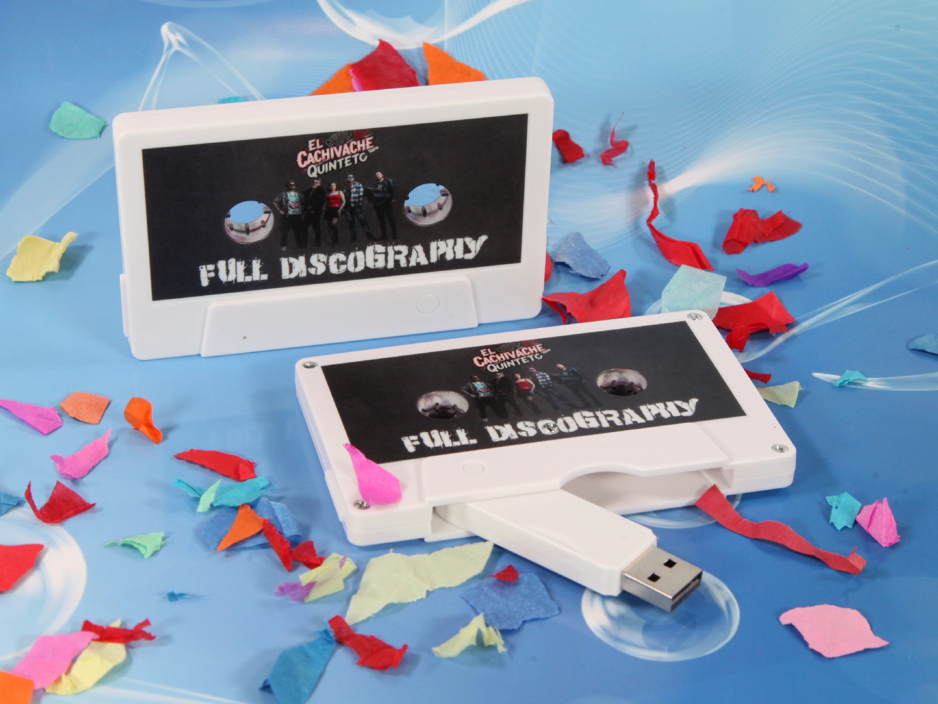 USB-Stick im Kassetten-Design mit individuellem Druckbereich und bunten Dekorationsstücken im Hintergrund.